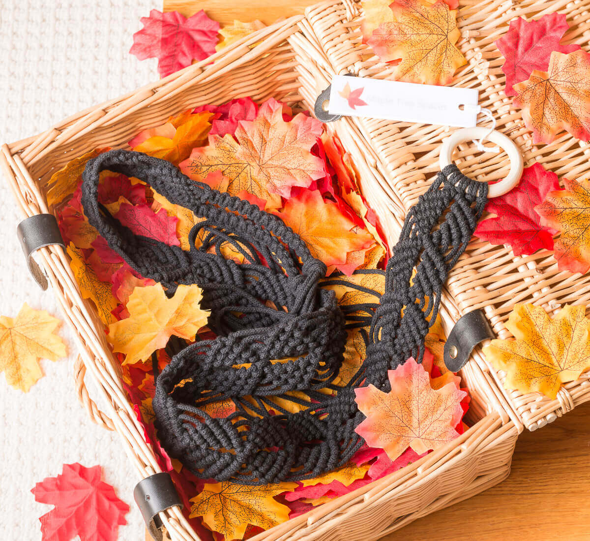 Black macrame plant hanger in a wicker basket surrounded by colorful autumn leaves.